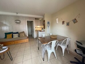 A living room with a sofa, a dining table with chairs, a kitchen in the background, and a tiled floor.