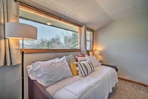 Un dormitorio con cama, lámpara de noche, ventana con persianas y vista a árboles.