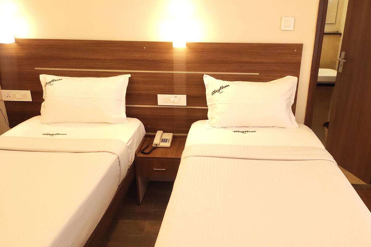 Ein Hotelzimmer mit zwei Betten, jedes mit einem 'Rhythm' beschrifteten Kopfkissen. Eine Nachttischlampe befindet sich auf der linken Seite des Raumes.