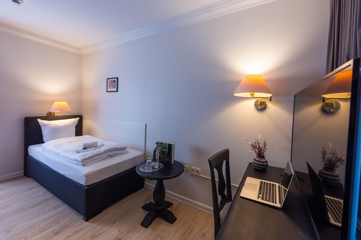 A hotel room with a bed, bedside table, two lamps, a desk with a laptop, and a mirror.