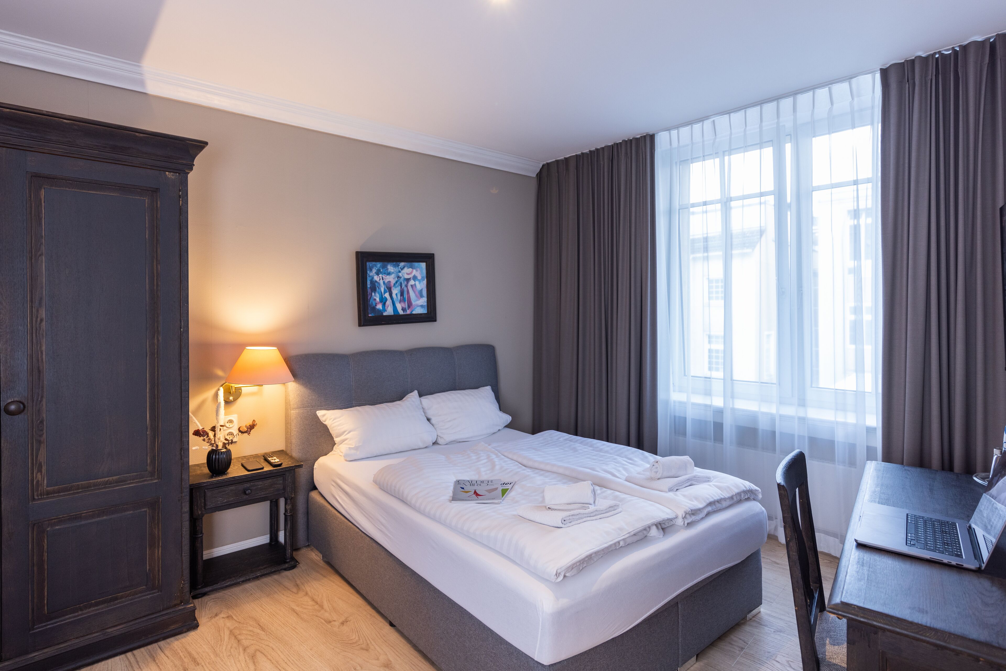 A hotel room with a bed, a desk with a laptop, a nightstand with a lamp, and a large window with curtains.