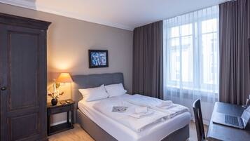 A hotel room with a bed, a desk with a laptop, a nightstand with a lamp, and a large window with curtains.