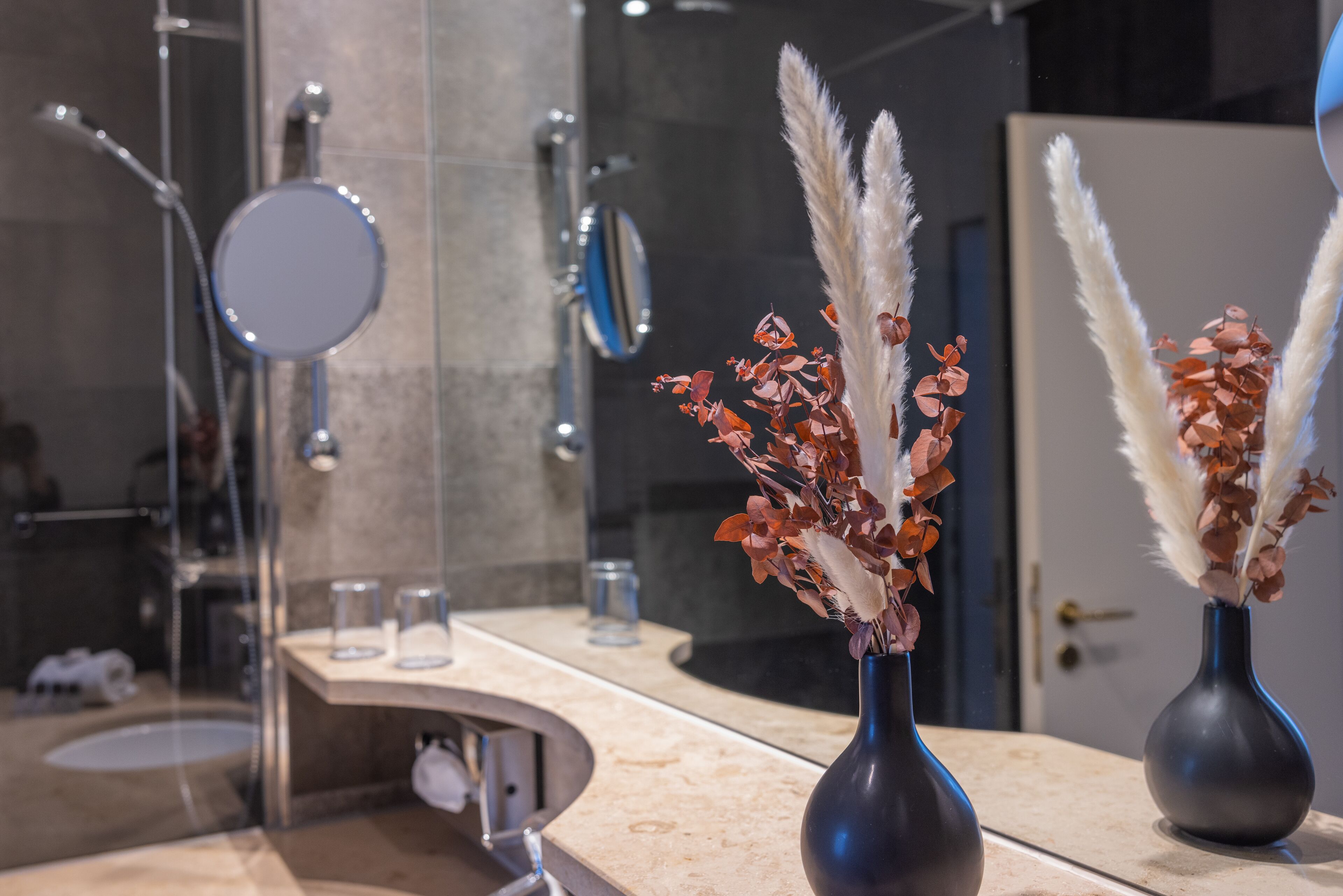 Une salle de bain avec un meuble-lavabo aux lignes courbes, deux vases foncés contenant des fleurs séchées, un miroir et un espace douche.