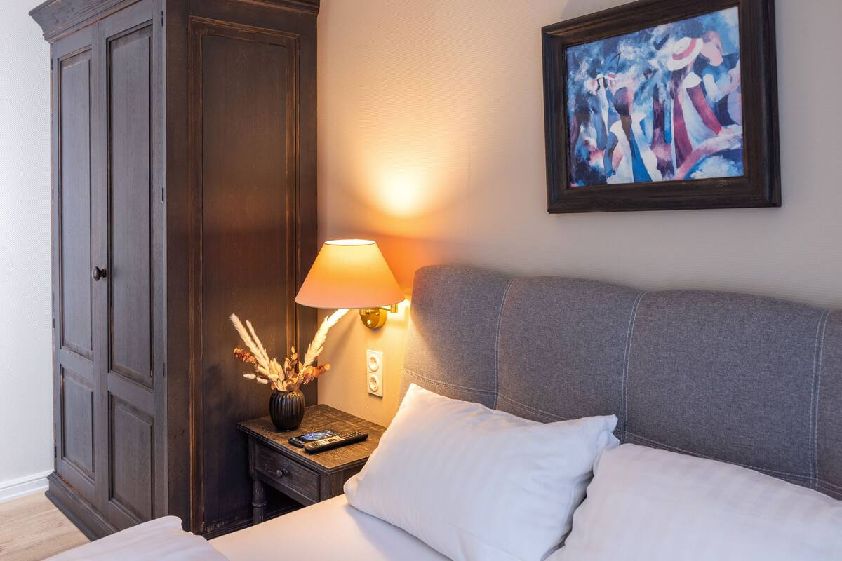 A hotel room with a bed, a nightstand with a lamp, a painting on the wall, and a wardrobe.