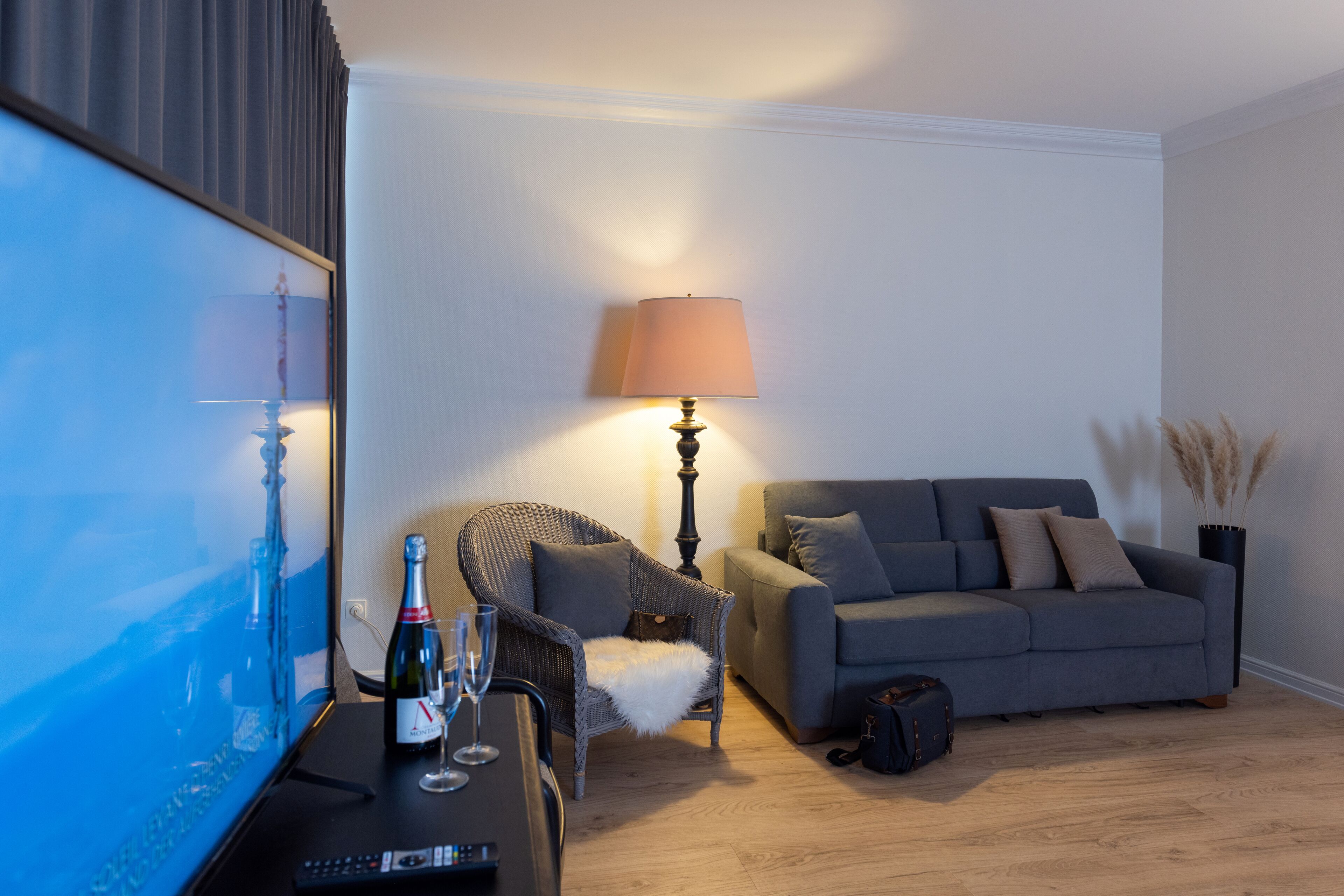 A living room with a sofa, a chair, a lamp, a TV, and a bottle of champagne on a table.