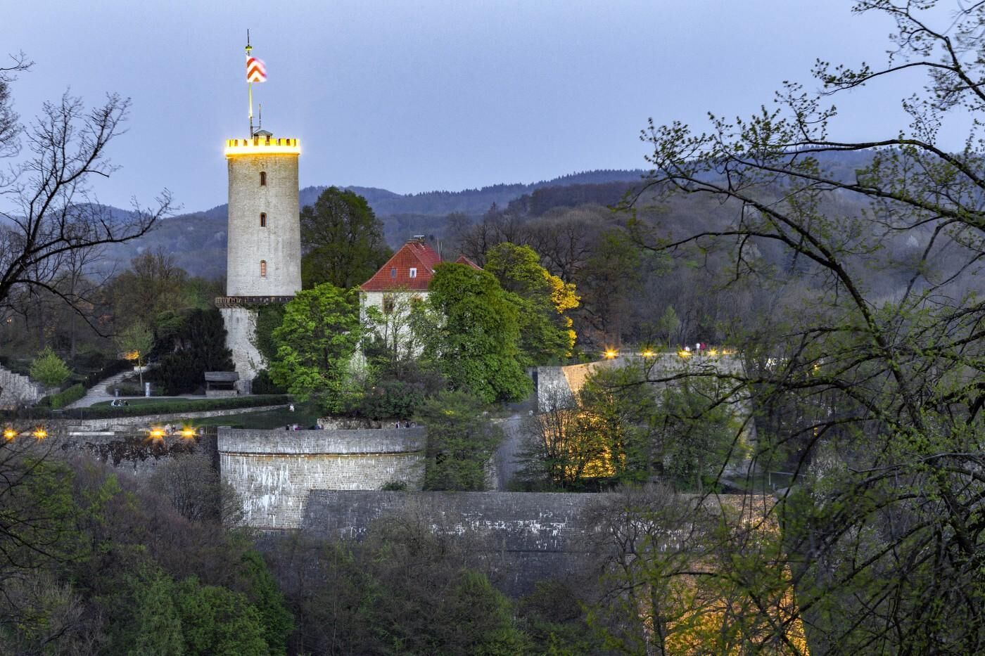 Una torre histórica con una bandera, rodeada de árboles y un muro de piedra.