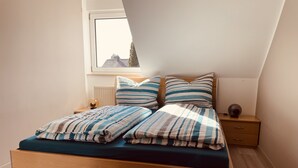 Quarto com cama de madeira, roupa de cama listrada, mesa de cabeceira, abajur e vista pela janela.