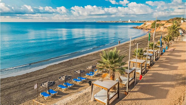 Una playa con sillas y sombrillas azules, un paseo marítimo arenoso y una vista despejada del mar.