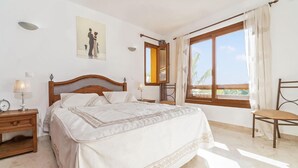 Un dormitorio con una cama grande, una mesita de noche, una silla, una ventana con cortinas y un cuadro en la pared.