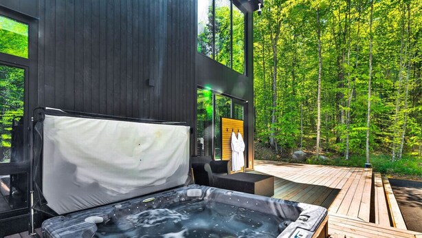 A modern outdoor hot tub area with a wooden deck, a large window overlooking a forest, and a cozy seating area. - Nordic Lodge Tremblant -5 Bdrs,Hot Tub,Pool Table (La Conception)