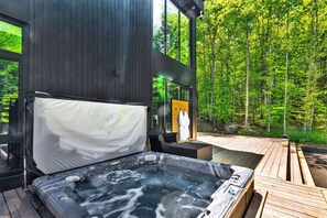 A modern outdoor hot tub area with a wooden deck, a large window overlooking a forest, and a cozy seating area. - Nordic Lodge Tremblant -5 Bdrs,Hot Tub,Pool Table (La Conception)