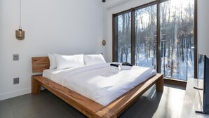 A modern bedroom with a wooden bed, white linens, and a large window offering a view of a snowy forest. - Nordic Lodge Tremblant -5 Bdrs,Hot Tub,Pool Table (La Conception)
