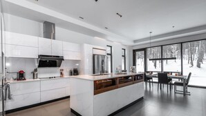 A modern kitchen with white cabinetry, a central island, and a dining area with a view of a snowy forest. - Nordic Lodge Tremblant -5 Bdrs,Hot Tub,Pool Table (La Conception)