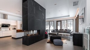A modern kitchen with white cabinets and a black marble accent wall, a living area with a fireplace, and large windows offering a view of the outdoors. - Nordic Lodge Tremblant -5 Bdrs,Hot Tub,Pool Table (La Conception)