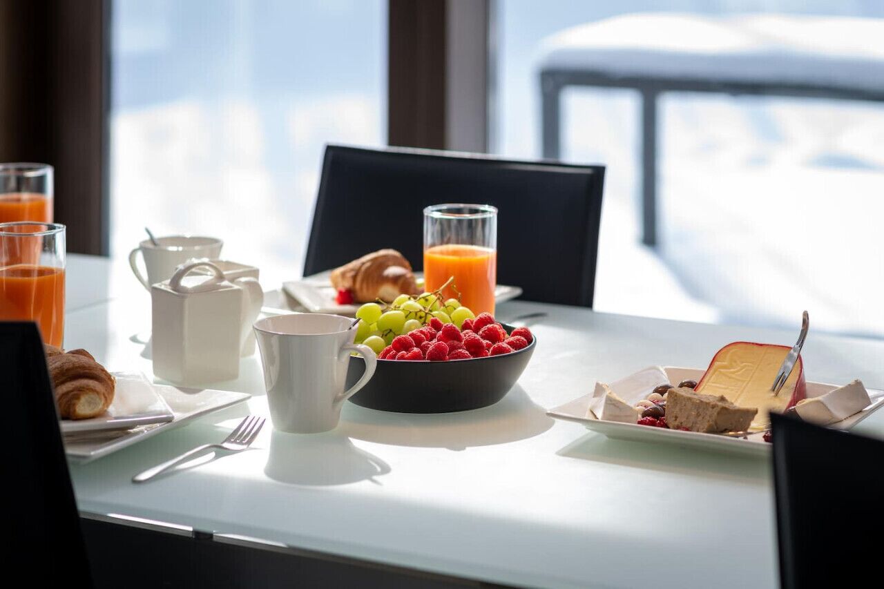 Een ontbijttafel met croissants, een fruitschaal, een glas sinaasappelsap en een bord met een plakje cake.