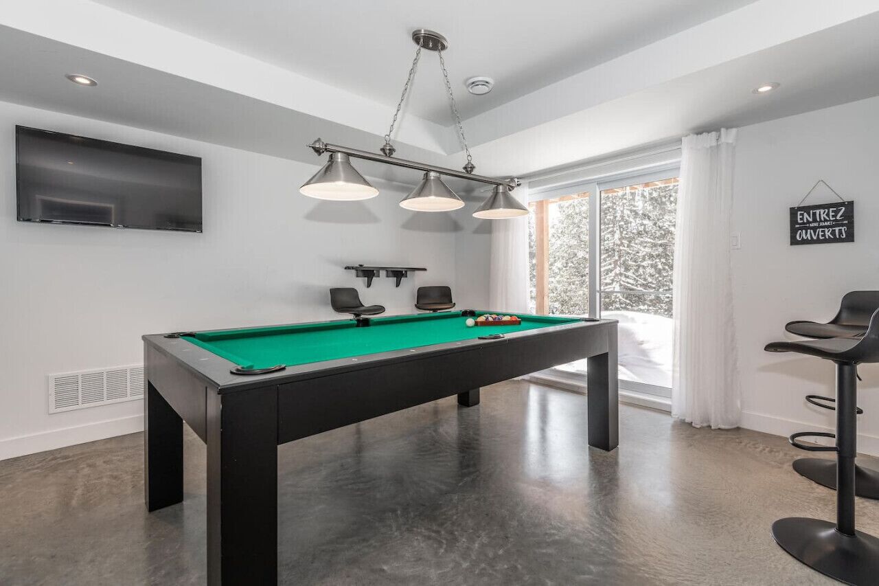 A modern room with a pool table, a mounted TV, and a large window with a view of trees.