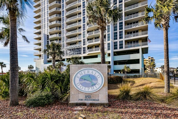 Une enseigne pour Emerald Dolphin, située au 850, Fort Pickens Road, avec un logo représentant un dauphin, entourée de palmiers et d’un bâtiment doté de plusieurs balcons.
