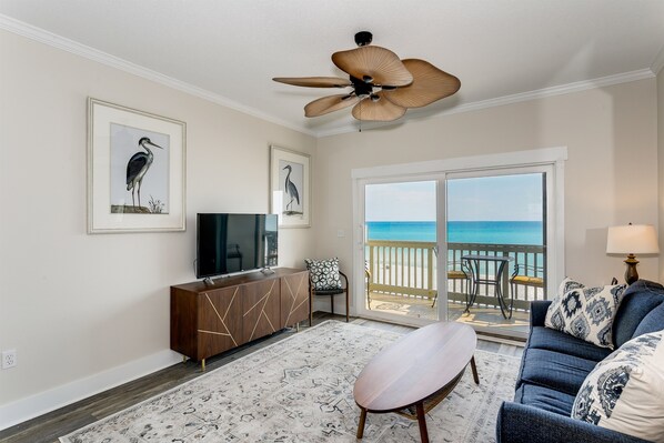 A living room with a large window, a ceiling fan, a TV, a sofa, and a view of the ocean.