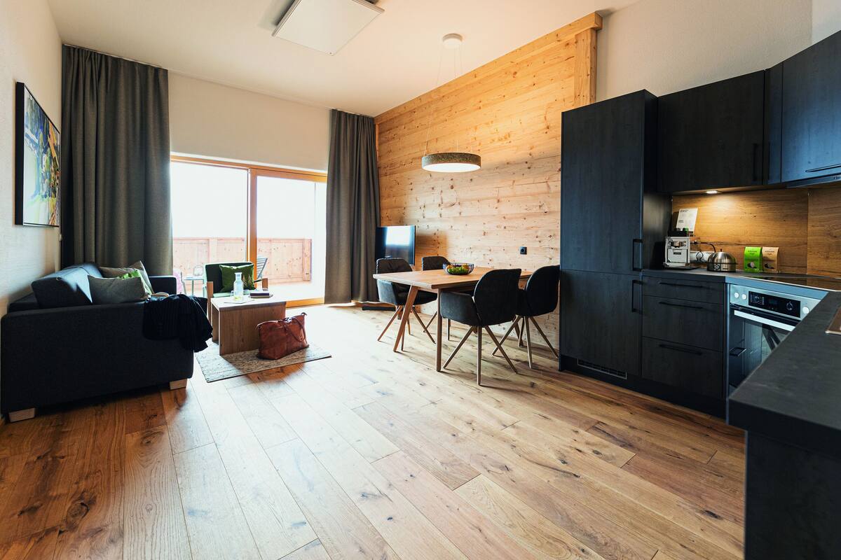 Ein modernes Wohnzimmer mit einer Couch, einem Esstisch und Stühlen. Ein Küchenbereich mit dunklen Schränken und Geräten ist sichtbar.