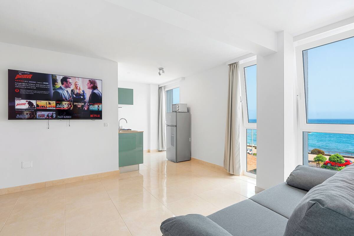 A modern living room with a large TV displaying movie posters, a kitchen area, and a view of the sea.
