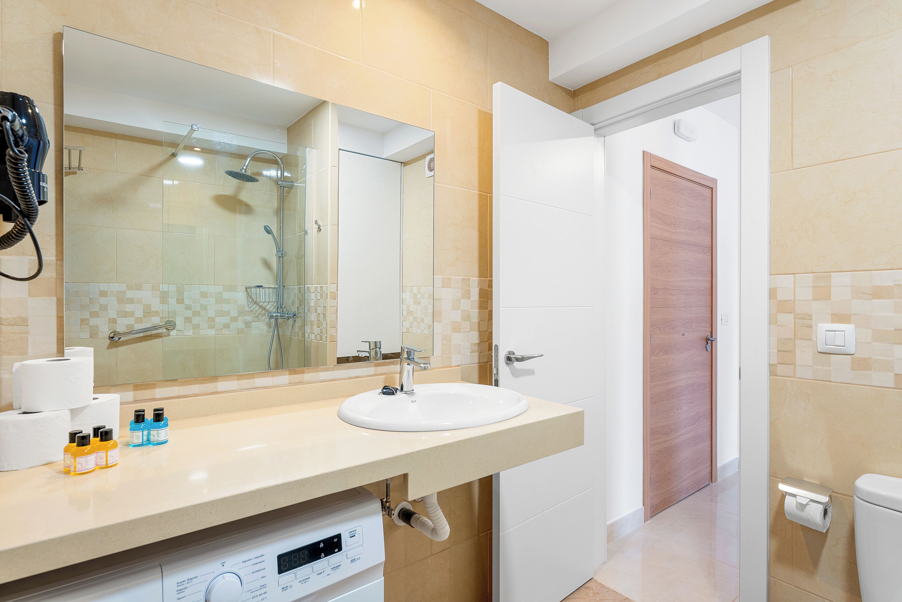 A bathroom with a sink, mirror, and shower. There are toiletries on the counter and a toilet visible in the background.