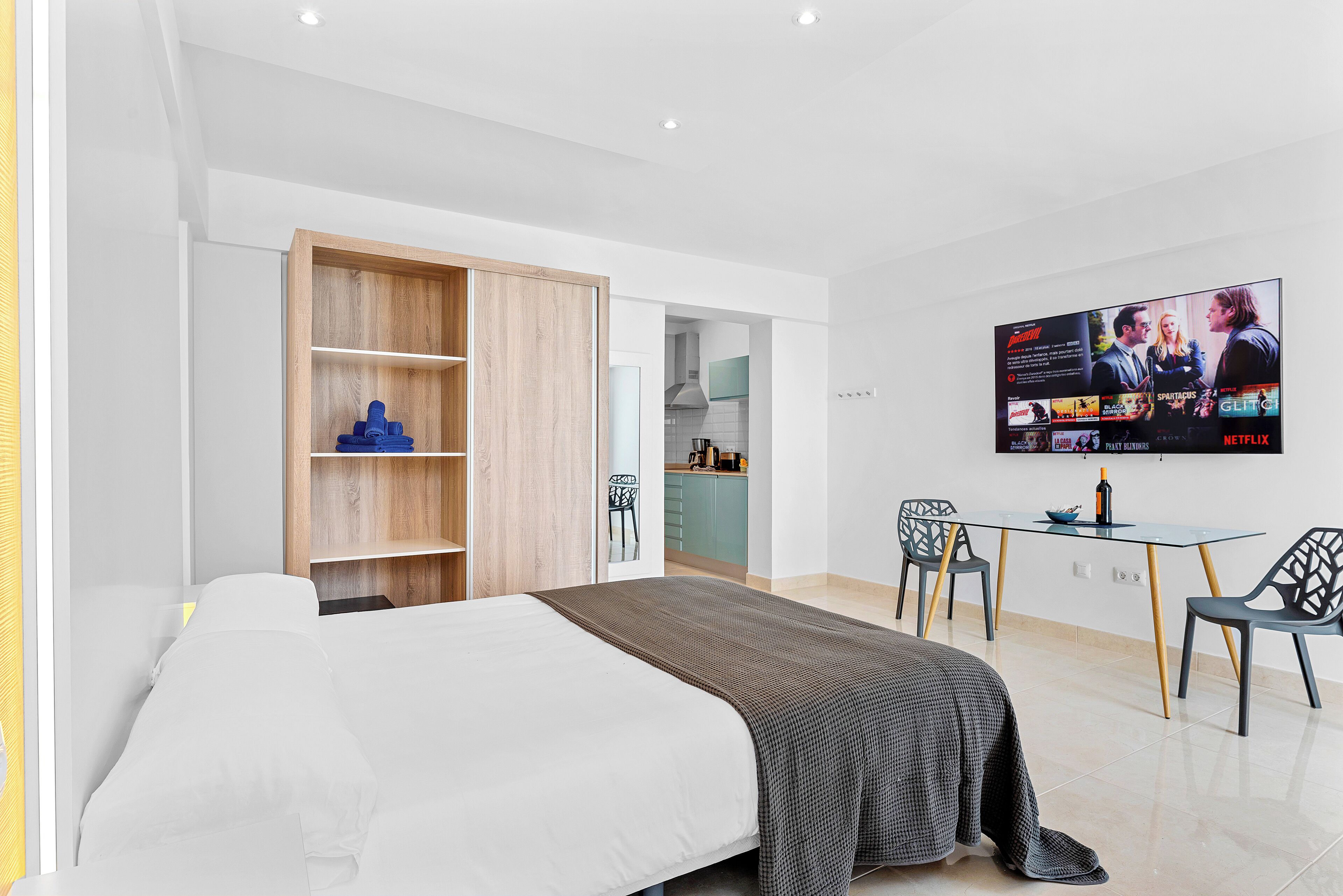 Une chambre moderne avec un lit, un téléviseur fixé au mur, une armoire en bois et un coin repas comprenant des chaises et une table.