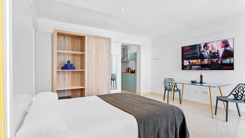 A modern bedroom with a bed, a TV mounted on the wall, a wooden wardrobe, and a dining area with chairs and a table.