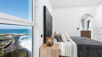 Une chambre avec une grande fenêtre donnant sur une plage et l’océan, un lit avec une tête de lit sombre et une table de chevet en bois surmontée d’une lampe décorative.