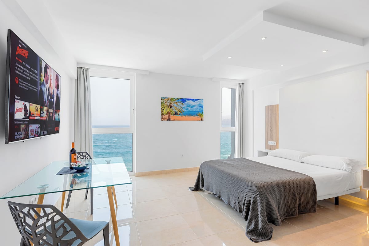 A modern hotel room with a large bed, a glass dining table, and a view of the sea.