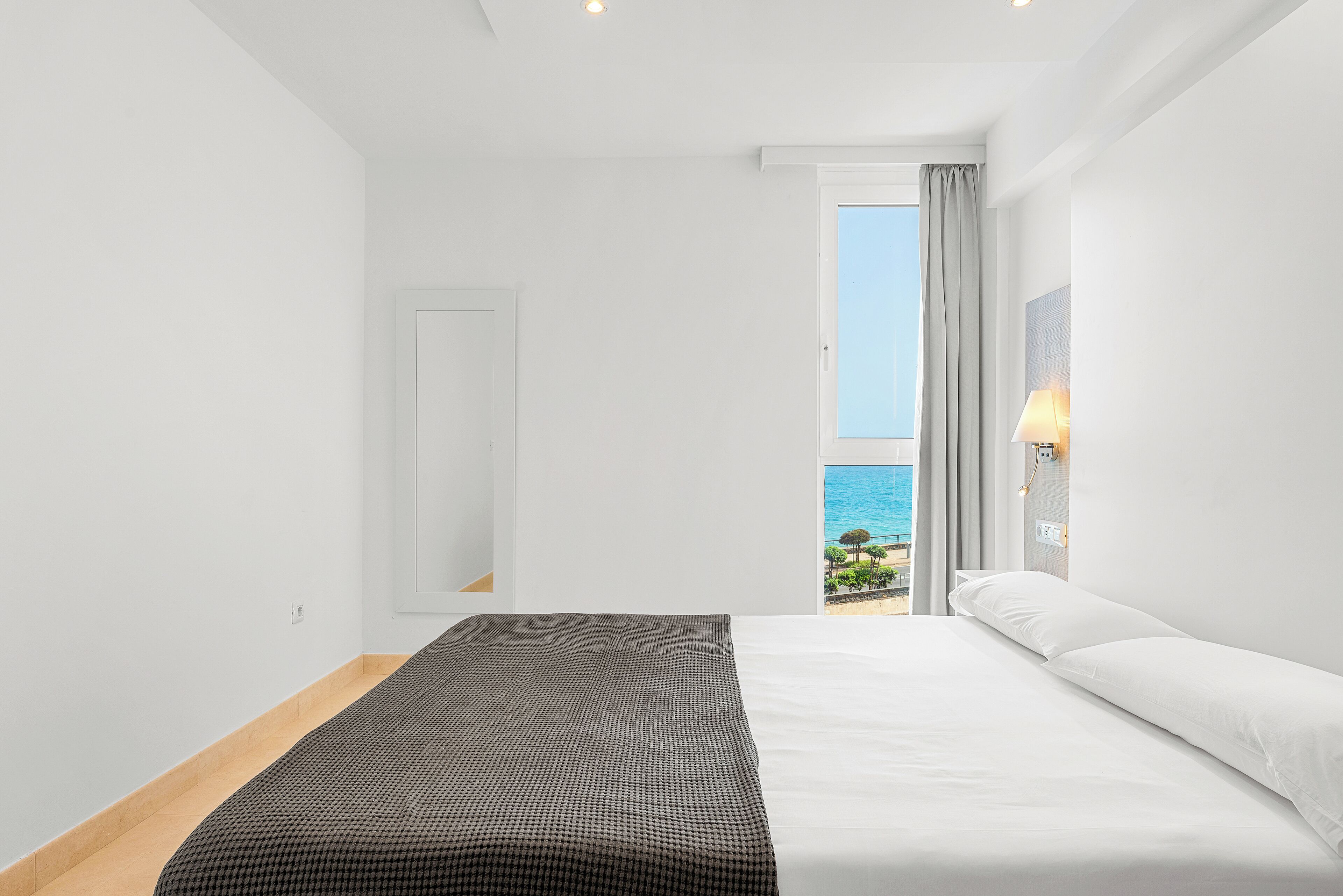 A hotel room with a bed, a window with a view of the sea, and a door.