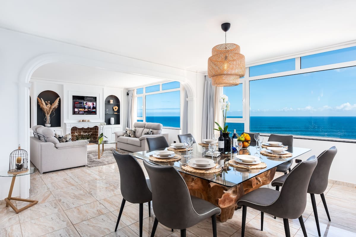 A modern dining area with a glass table, dark chairs, and a view of the ocean.