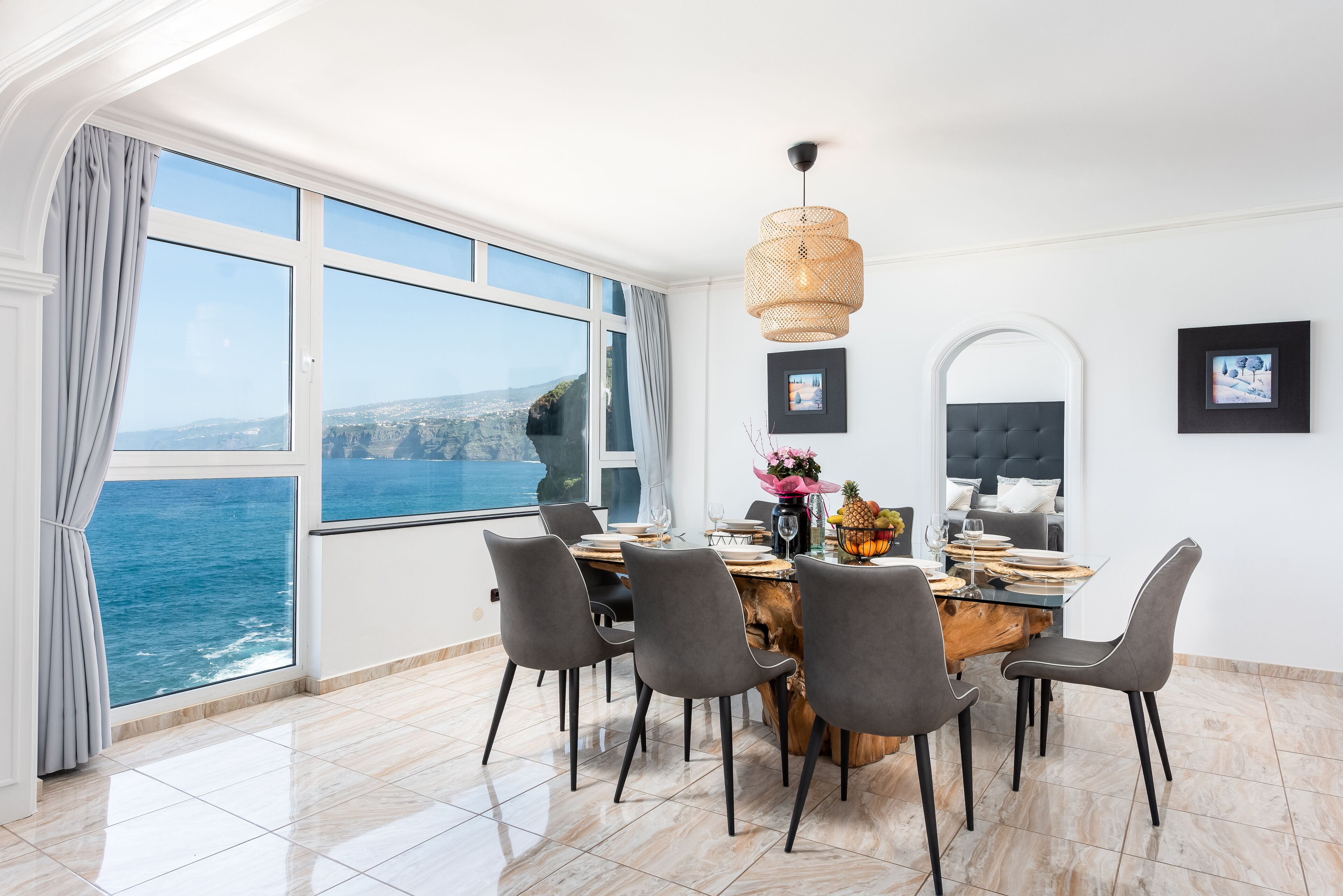 Un comedor con un amplio ventanal con vistas al mar, una mesa redonda de madera y sillas grises.