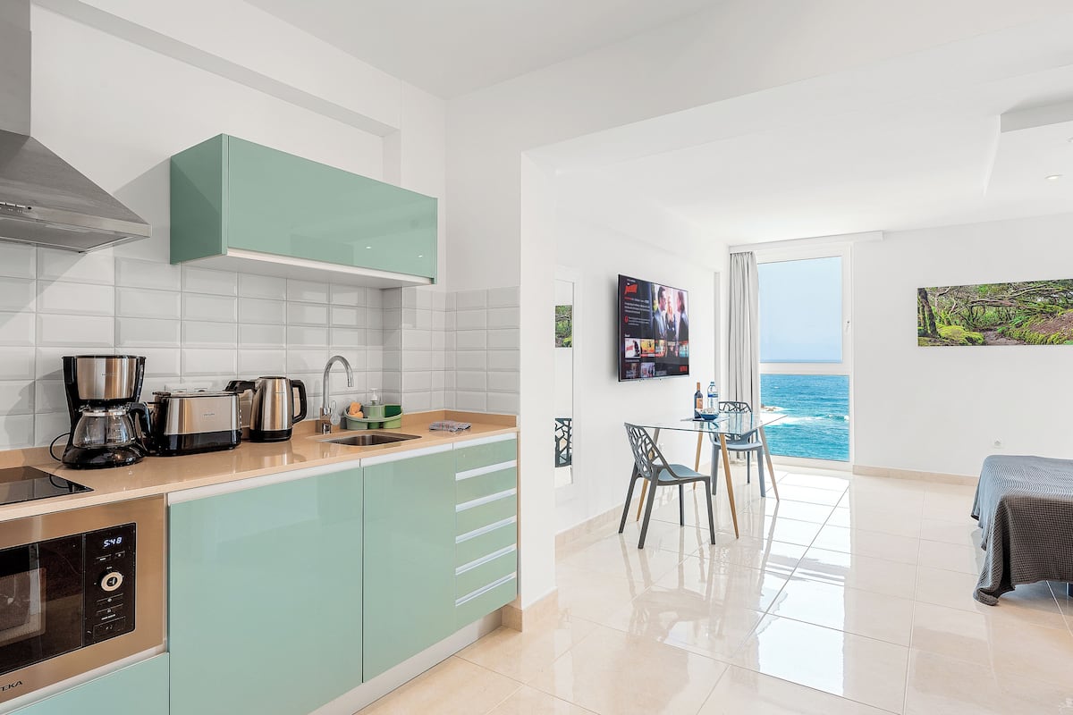 A modern kitchen with a view of the sea, a dining area with a table and chairs, and a bed in the adjacent room.