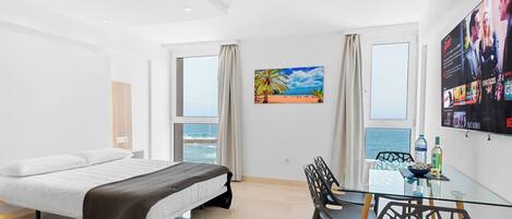 Una habitación de hotel moderna con una cama, una mesa de comedor de vidrio, sillas, un televisor y un cuadro con una escena de playa.