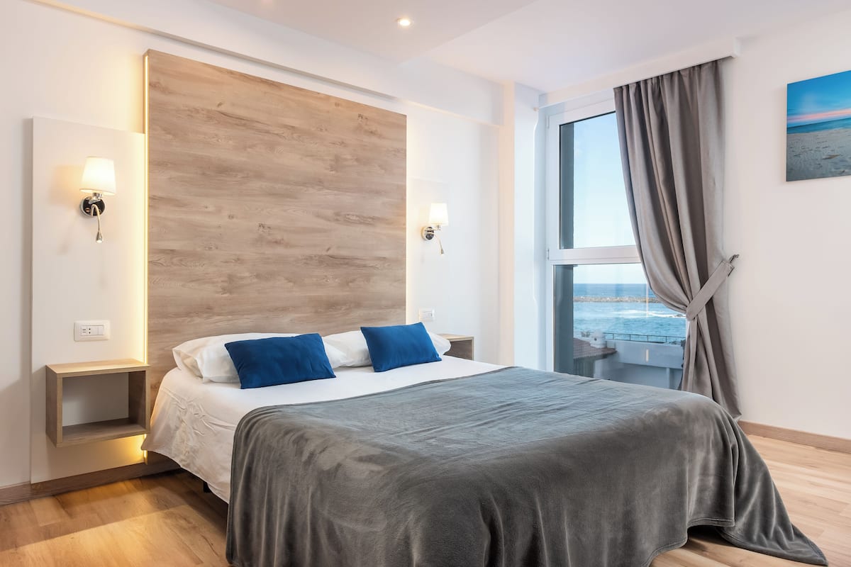 A modern hotel room with a large bed, wooden headboard, bedside lamps, and a view of the sea through a large window.
