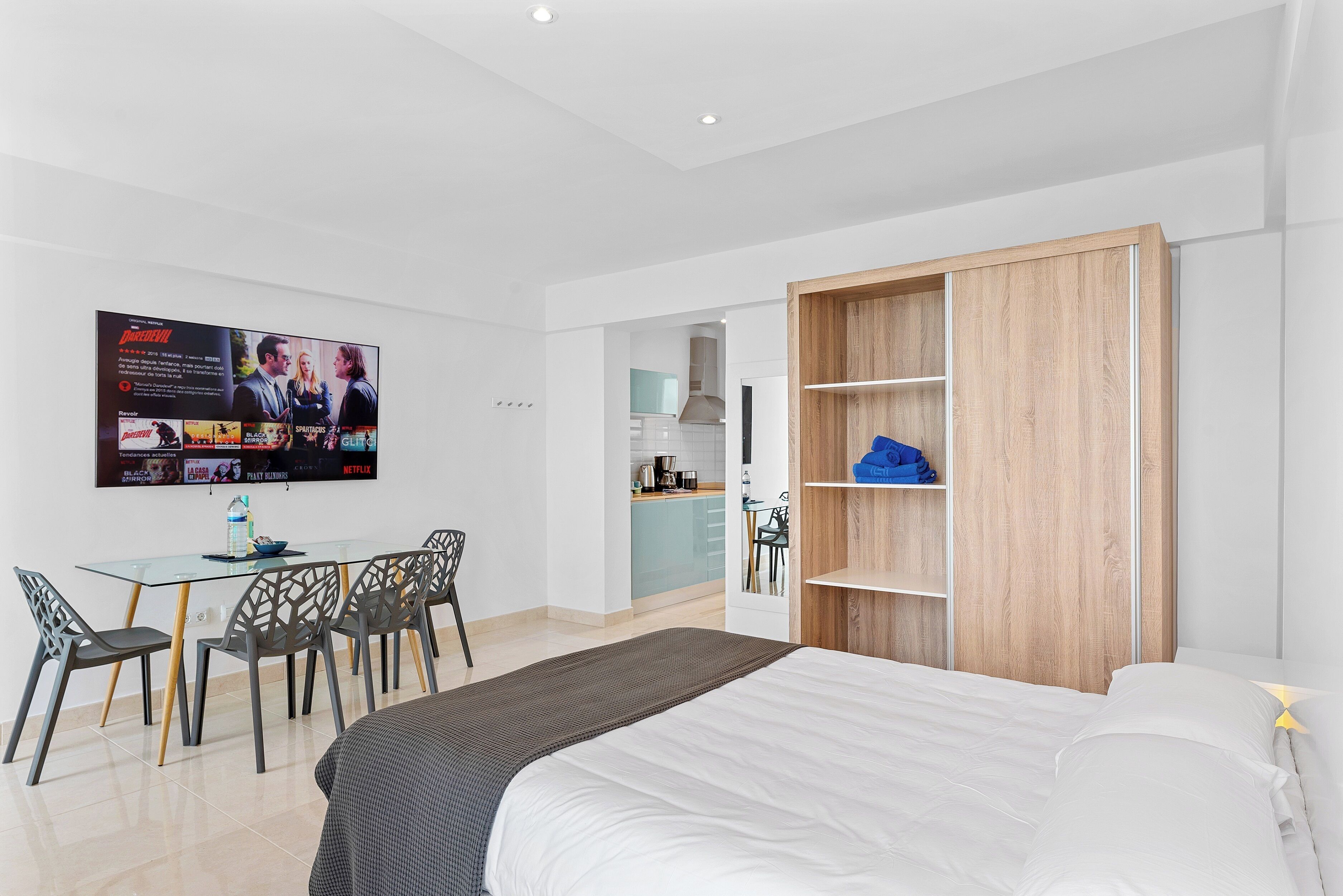A modern hotel room with a bed, a TV mounted on the wall, a dining area with chairs, and a kitchenette in the background.