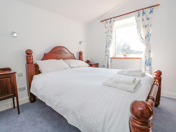 A neatly made bed with white linens and a wooden headboard, a bedside table with a lamp, and a window with curtains.