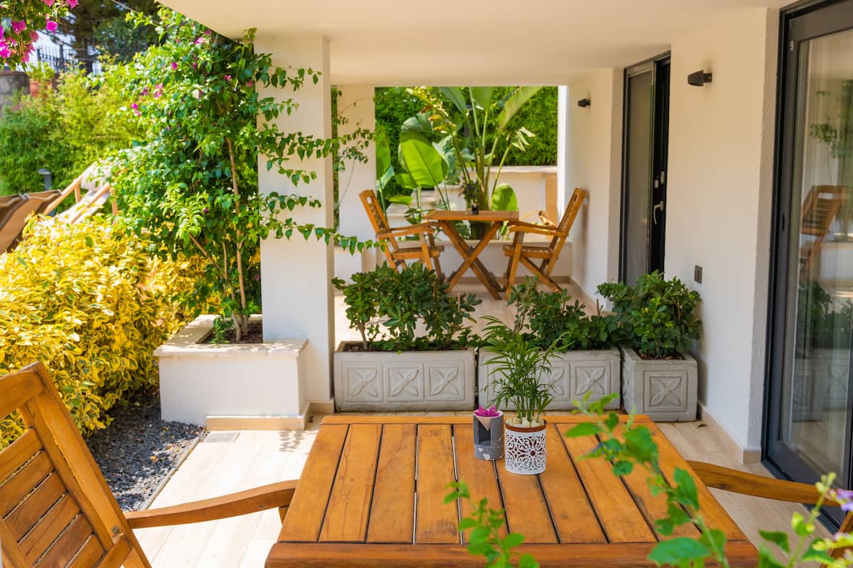 An outdoor patio with a wooden table and chairs, surrounded by potted plants and a variety of greenery.
