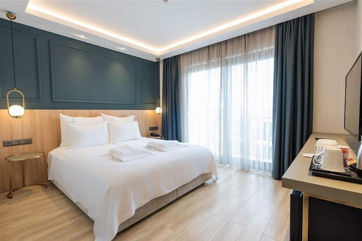 A hotel room with a large bed, bedside tables, a desk with a chair, and a large window with curtains.