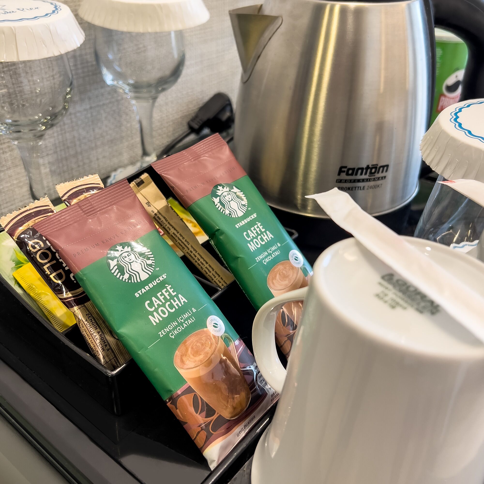 A tray with Starbucks Caffe Mocha and Gold packets, a white mug, and a stainless steel pot.