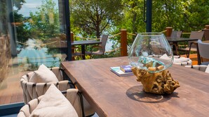 A wooden table with a glass vase and a book, surrounded by cushioned chairs, with a view of greenery and a rope barrier. - Cunda Pier Otel (Ayvalik)