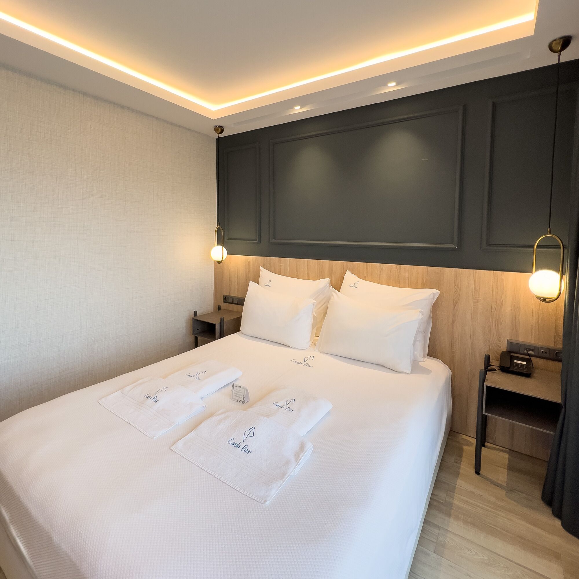 A neatly made bed with white linens and towels, a wooden headboard, bedside tables with lamps, and a modern ceiling light fixture.