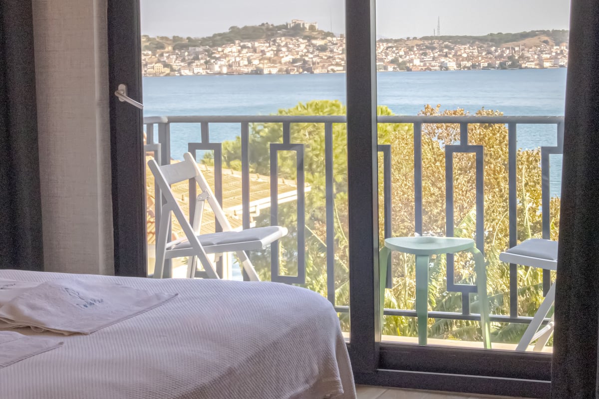 A balcony with a bed and chairs, overlooking a coastal cityscape.