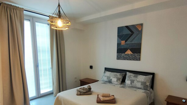 A modern bedroom with a large bed, a hanging light fixture, a painting on the wall, and a view through the window. - Central apartment in private area (Barletta)