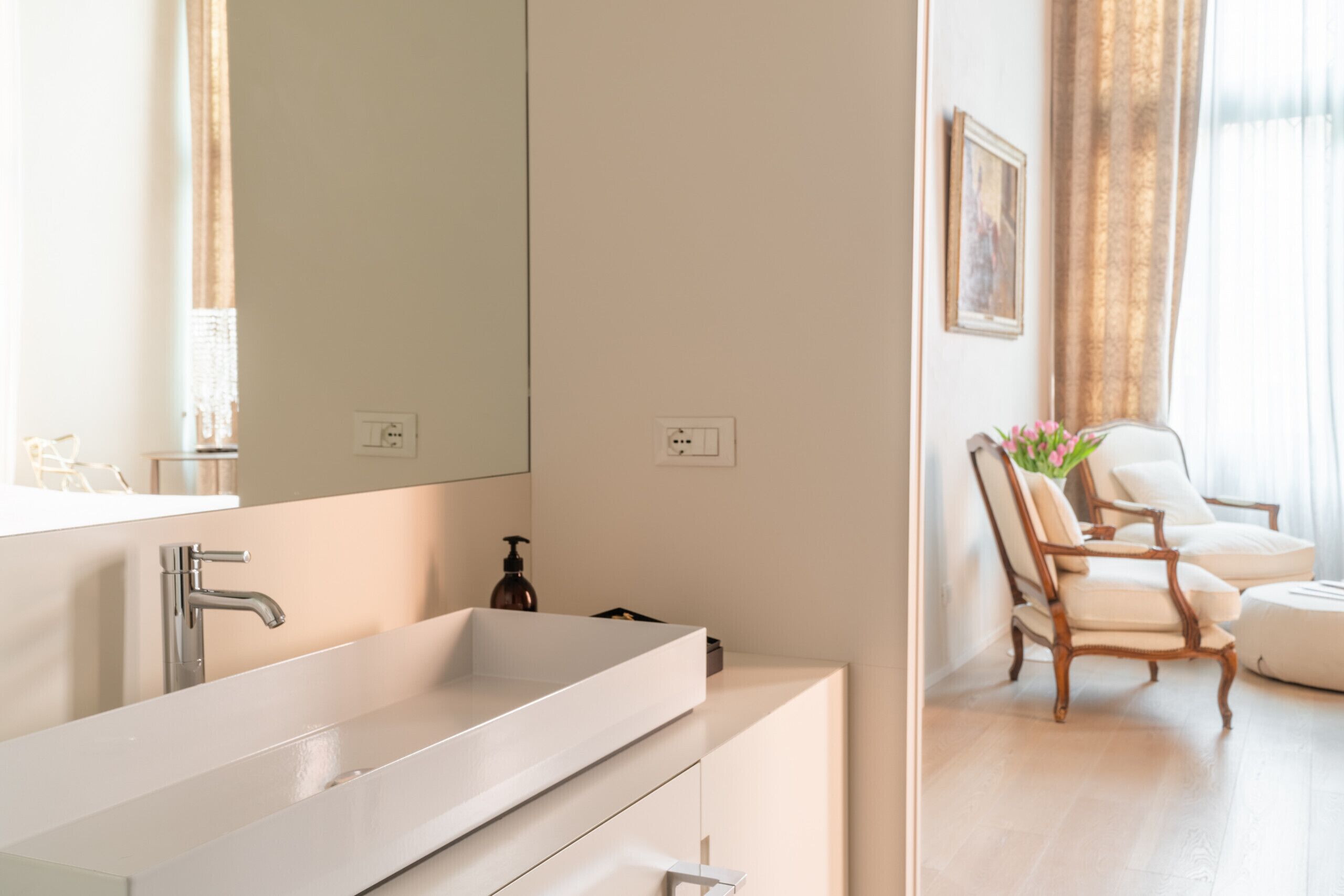 Un bagno moderno con un ampio lavandino bianco, uno specchio e una sedia di legno sullo sfondo.