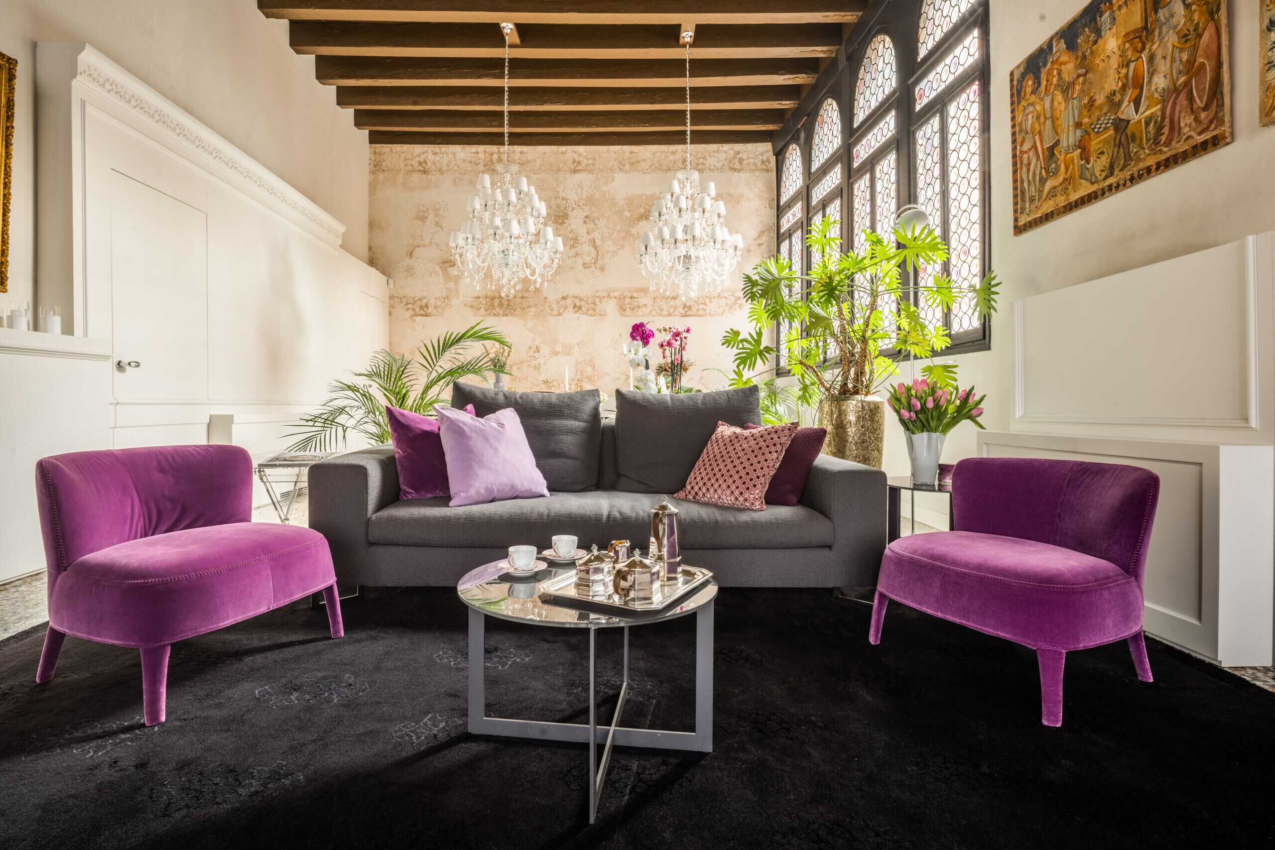 Ein Wohnzimmer mit einem grauen Sofa, zwei violetten Sesseln, einem Couchtisch und einem schwarzen Teppich. Es gibt zwei Kronleuchter, ein Fenster mit Pflanzen und ein Gemälde an der Wand.