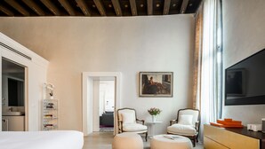 Ein modernes Hotelzimmer mit einem großen Bett, zwei weißen Sesseln, einem kleinen runden Tisch, einem Fernseher und einem Bücherregal.