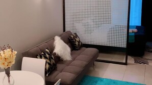 A modern living room with a sofa, a dining table with chairs, a blue rug, and a ceiling light fixture. - Stay.Plus Royal Suburbs By Tsavo (Nairobi)