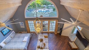 A loft-style room with a high ceiling, a large arched window, a ceiling fan, a hanging light fixture, and a cozy seating area with a patterned rug and cushions. - Spacious 5BR Helen villa with private pool, hot tub, and game room. (Helen)