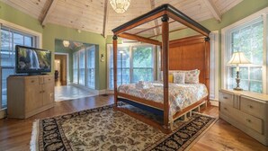 A four-poster bed with a floral bedspread, a wooden nightstand, a television on a cabinet, and a large window with blinds. - Spacious 5BR Helen villa with private pool, hot tub, and game room. (Helen)